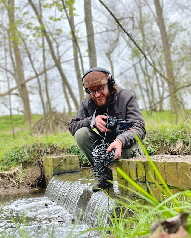 Gregory White in a forest recording a running river with a hydrophone. His face is lit up with happiness and joy at the sounds he is hearing!