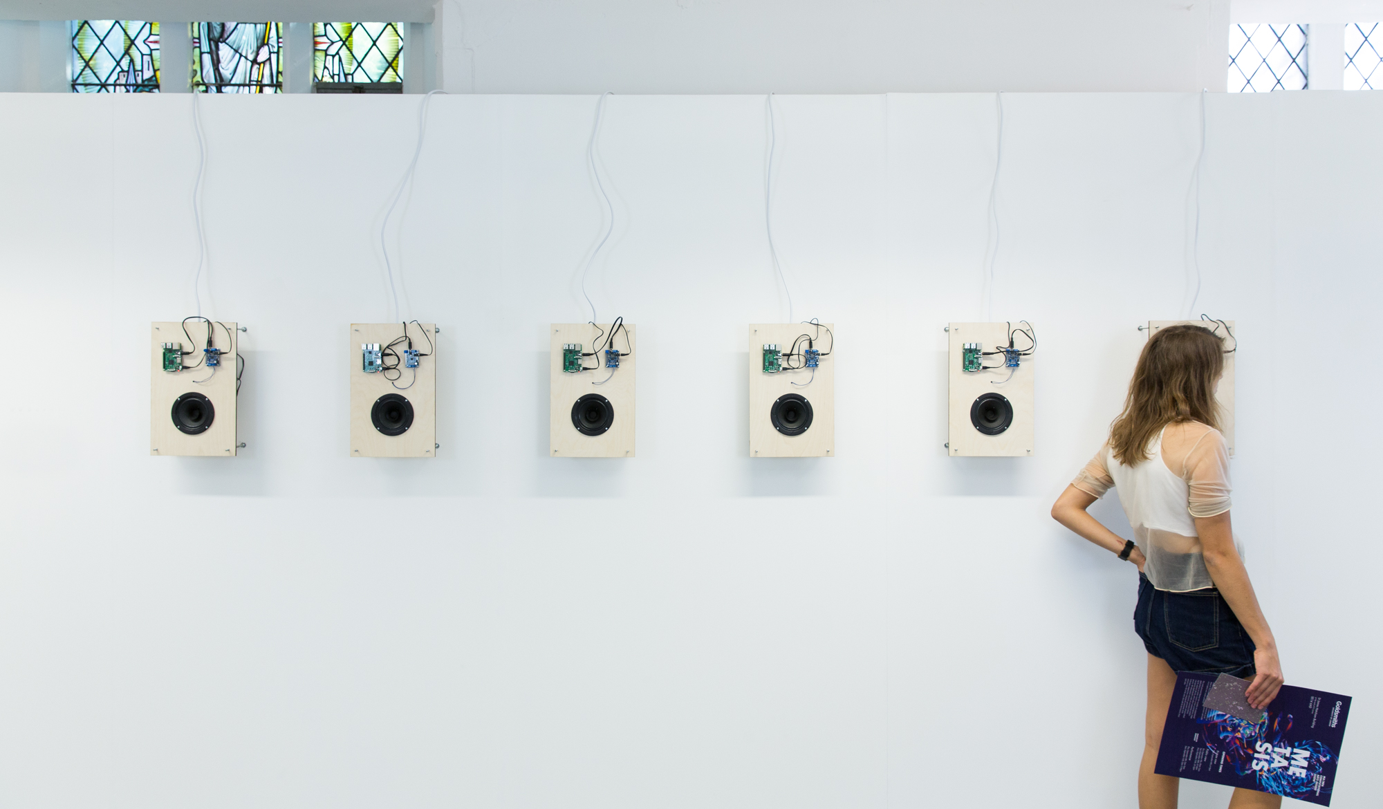 Six wooden boards are mounted to a white gallery wall, each with electronics and a speaker cone attached. A woman listens intently by one of the speaker cones.